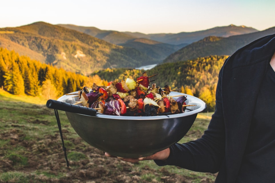 Hånd som holder en bolle med grillspyd på en bakketopp med skog i bakgrunnen