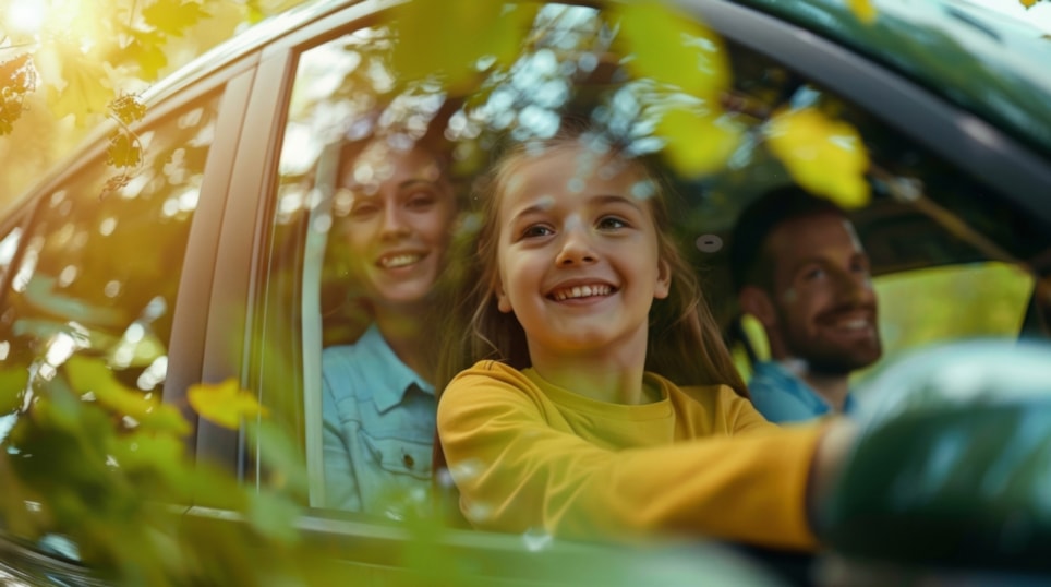 Familie i bil, jente i forgrunnen som ser ut av bil smilende